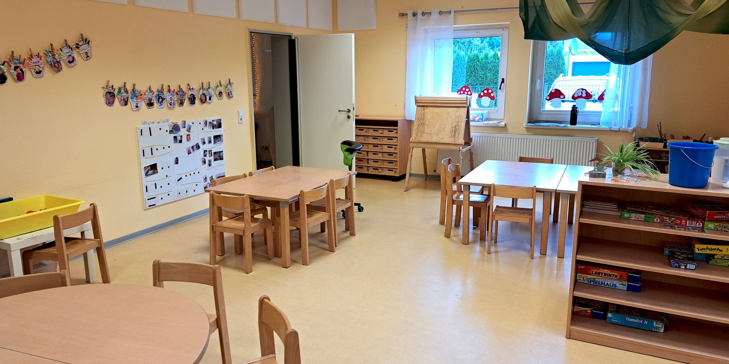 Heller Gruppenraum der Kita Villa Kunterbunt mit kleinen Holztischen und -stühlen, einem Regal mit Spielen, einer Staffelei am Fenster, aufgehängten Kinderarbeiten an der Wand und einem grünen Stoffdach über einem Tisch.
