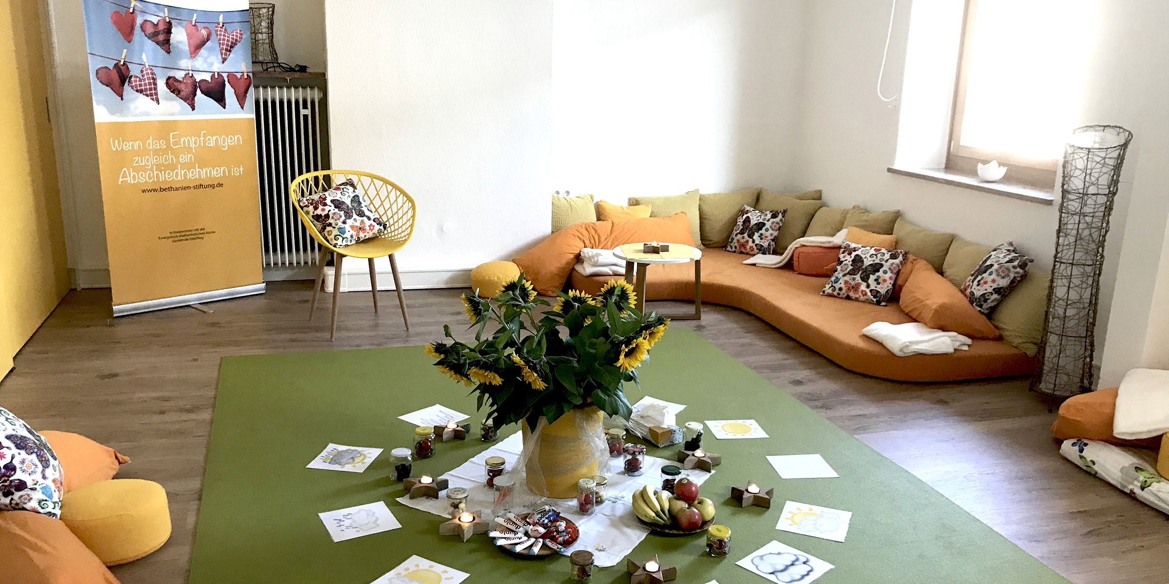 Ein gemütlichen Raum der Einrichtung mit einem Sofa und vielen Kissen. In der Mitte des Raumes ist ein großer Sonnenblumenstrauß platziert. 