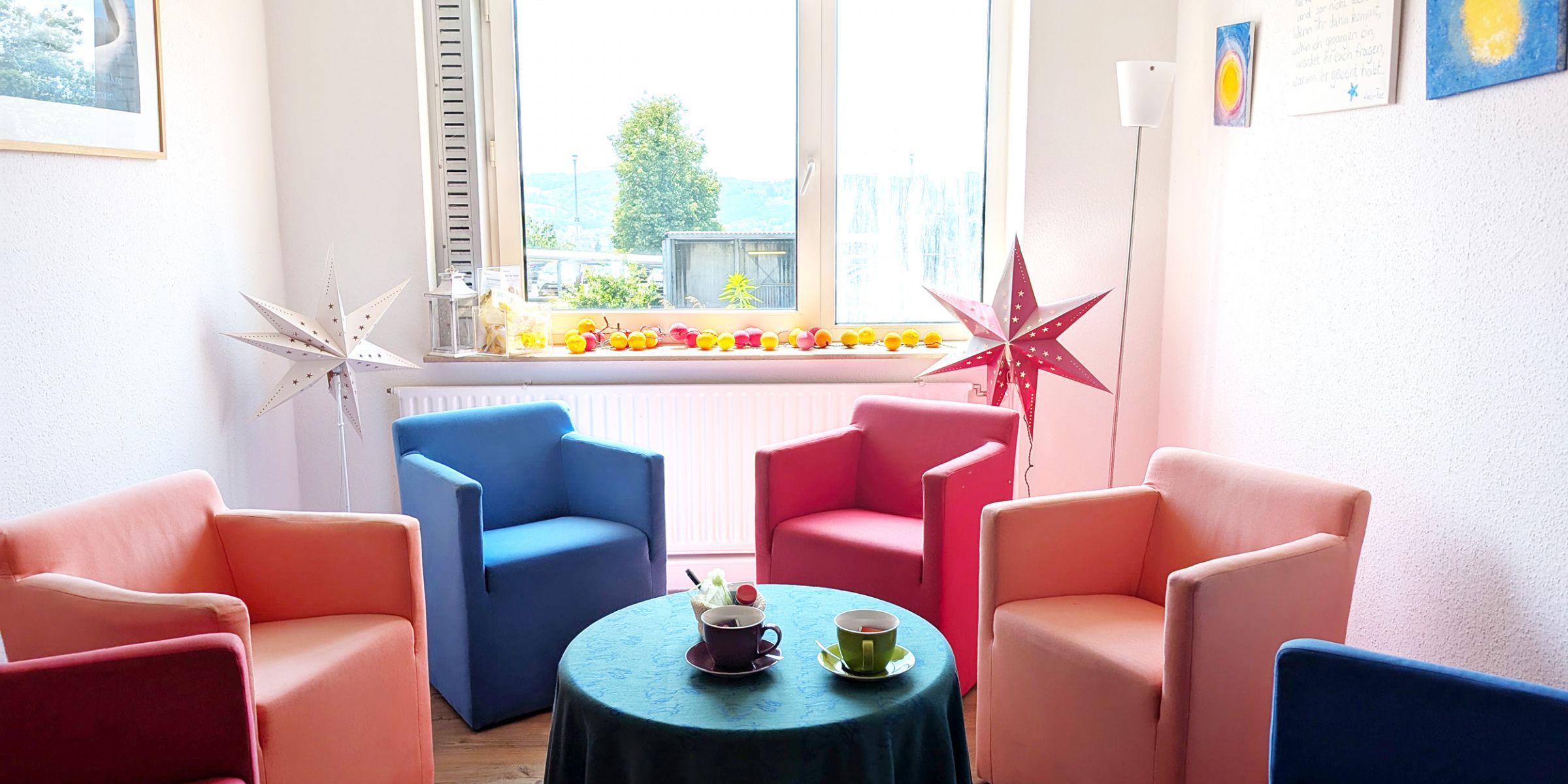 Das Bild zeigt einen Raum in denen Sessel in unterschiedlichen Farben in einem Kreis stehen. In der Mitte steht ein Tisch mit zwei Tassen und hinter den Sesseln stehen zwei Stern Lampen.