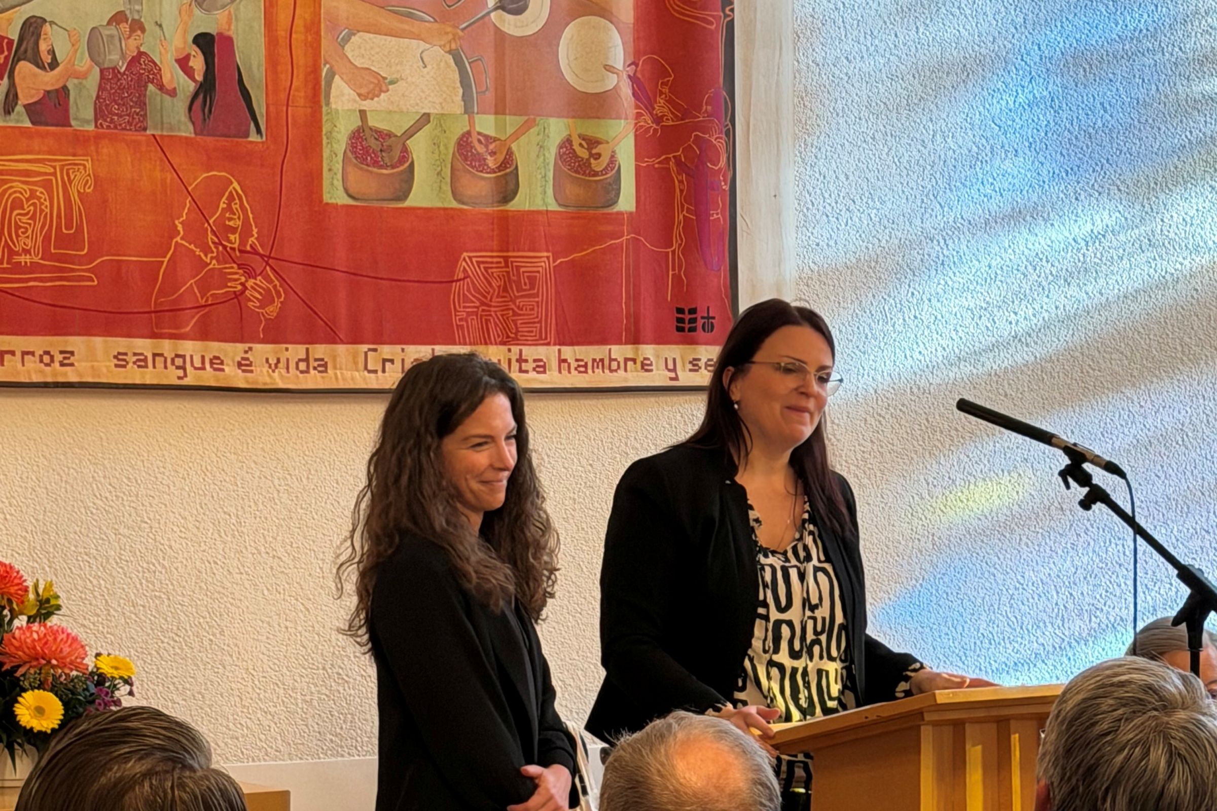 Ramona Schulze und Stefanie Wittkowski halten gemeinsam ihre Ansprache im hellen Altarraum vor einem orange-rötlichen Wandbild