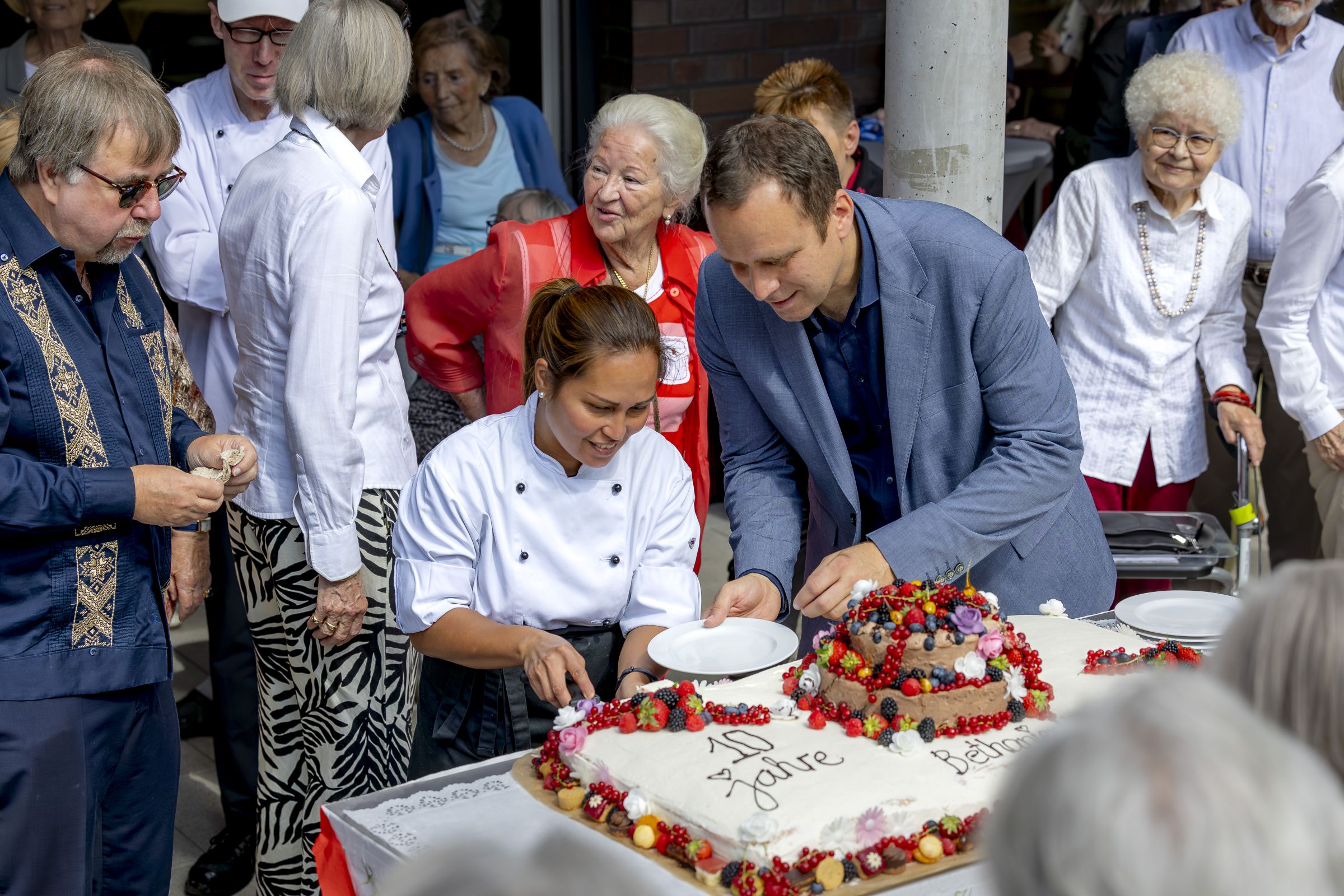 Eine große rechteckige Torte mit weißer Creme und vielen frischen Früchten wird angeschnitten
