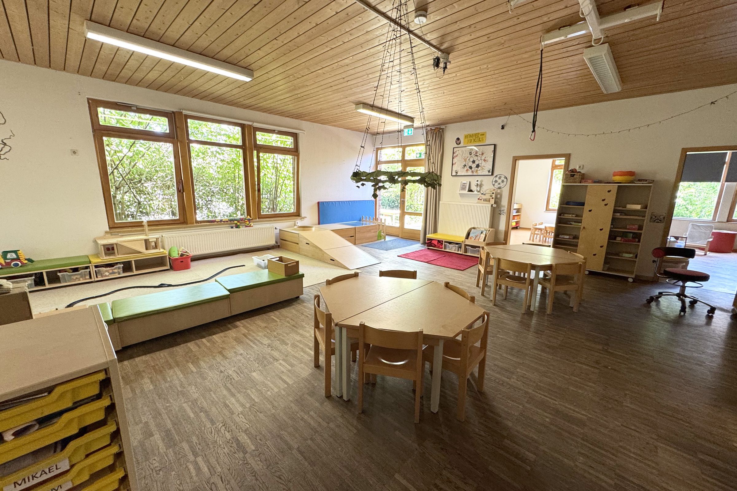 Ein Gruppenraum der Kita Arche Kunterbunt in Monheim mit Holzboden, kleinen Tischen und Stühlen und viel Platz zum Spielen