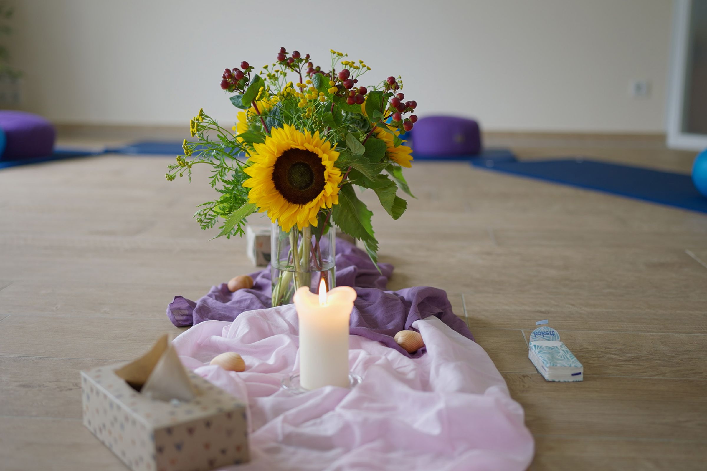 Ein Blumenstrauß auf dem Boden, vor dem eine brennende Kerze steht mit Taschentüchern und Muscheln. Der Raum darum ist verschwommen und ähnelt einem Yoga-Raum mit Matten.