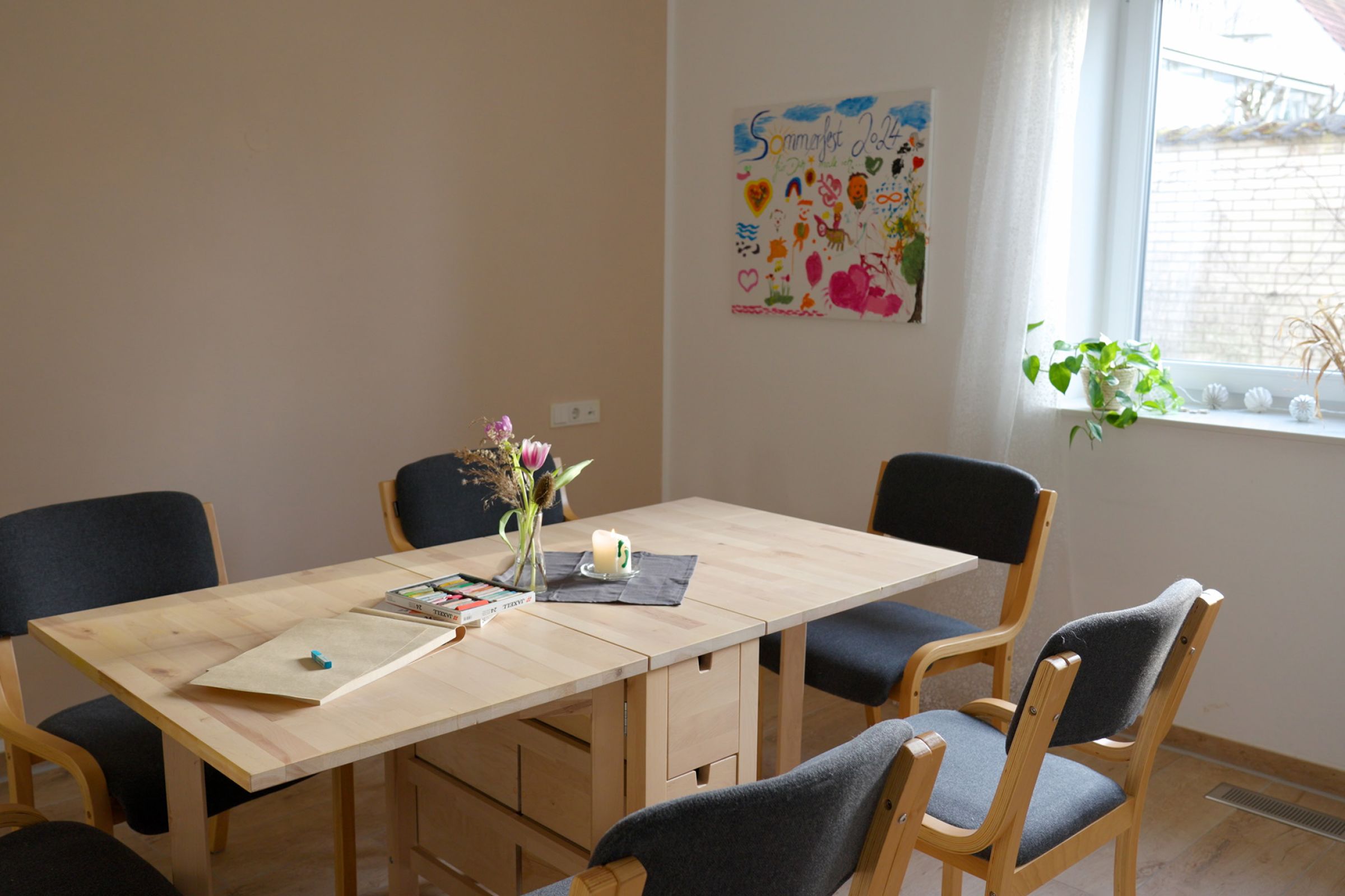 Ein Raum der Einrichtungen in dem ein hölzerner Tisch steht auf dem sich Malkreide, sowie Blumen und eine kleine Kerze befinden. Der Raum wirkt offen und einladend.