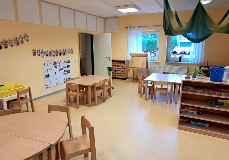 Heller Gruppenraum der Kita Villa Kunterbunt mit kleinen Holztischen und -stühlen, einem Regal mit Spielen, einer Staffelei am Fenster, aufgehängten Kinderarbeiten an der Wand und einem grünen Stoffdach über einem Tisch.