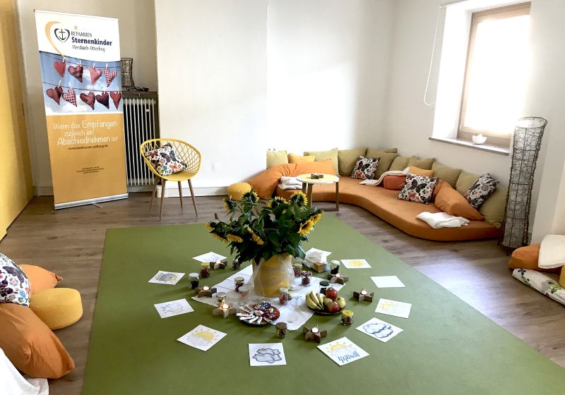 Ein gemütlichen Raum der Einrichtung mit einem Sofa und vielen Kissen. In der Mitte des Raumes ist ein großer Sonnenblumenstrauß platziert. 