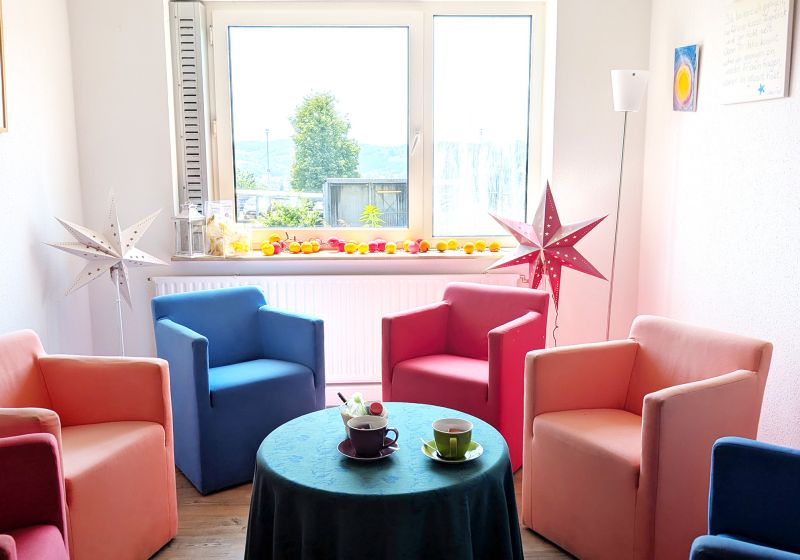 Das Bild zeigt einen Raum in denen Sessel in unterschiedlichen Farben in einem Kreis stehen. In der Mitte steht ein Tisch mit zwei Tassen und hinter den Sesseln stehen zwei Stern Lampen.